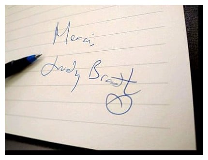 Handwritten thank-you note reading “Merci, Judy Bradt” in blue ink on lined paper with a pen visible